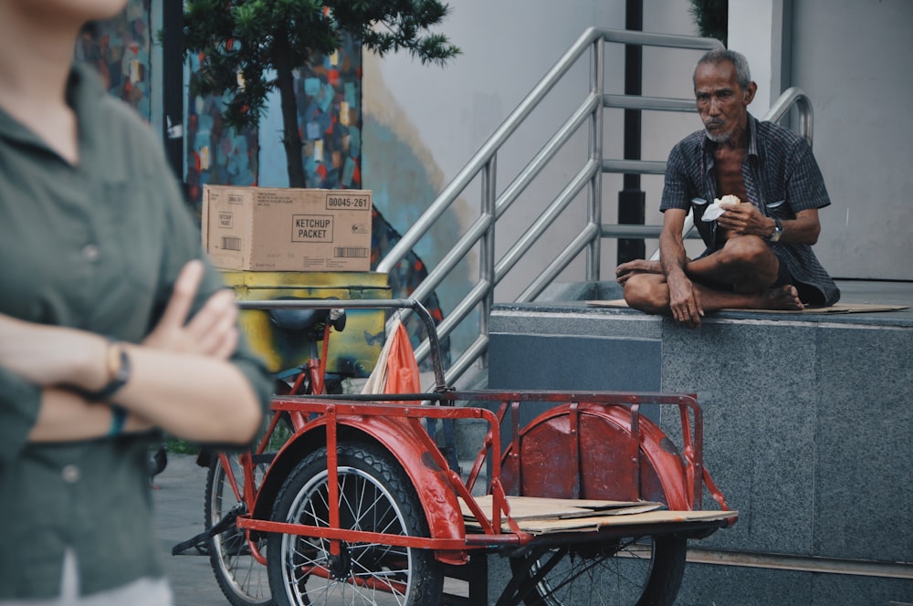 man sitting on curb in front of red trike at daytime