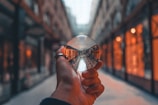 A hand is holding a clear crystal ball in an indoor shopping arcade. The ball captures an inverted reflection of the corridor, showcasing a row of warmly lit shop windows lining the path. The scene has a warm, cozy ambiance with soft lighting creating a sense of depth.