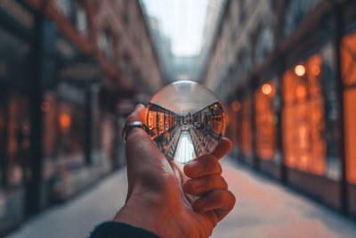 A hand is holding a clear crystal ball in an indoor shopping arcade. The ball captures an inverted reflection of the corridor, showcasing a row of warmly lit shop windows lining the path. The scene has a warm, cozy ambiance with soft lighting creating a sense of depth.