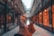 A hand is holding a clear crystal ball in an indoor shopping arcade. The ball captures an inverted reflection of the corridor, showcasing a row of warmly lit shop windows lining the path. The scene has a warm, cozy ambiance with soft lighting creating a sense of depth.