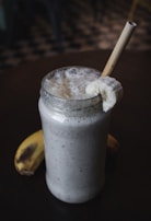 A creamy banana smoothie served in a glass jar with frothy texture on top. A slice of banana is placed on the rim of the jar, and a bamboo straw is inserted. Another banana lies on the dark surface beside the jar.