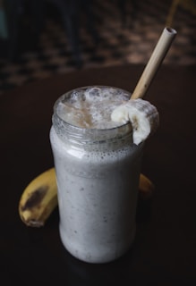 A creamy banana smoothie served in a glass jar with frothy texture on top. A slice of banana is placed on the rim of the jar, and a bamboo straw is inserted. Another banana lies on the dark surface beside the jar.