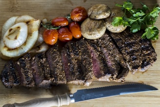 A beautifully arranged plate of sliced grilled steak on a wooden board accompanied by grilled onions, sautéed mushrooms, roasted cherry tomatoes, and fresh parsley. A serrated knife is placed nearby, adding to the rustic presentation.