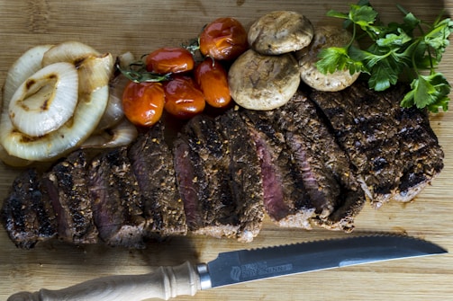 A beautifully arranged plate of sliced grilled steak on a wooden board accompanied by grilled onions, sautéed mushrooms, roasted cherry tomatoes, and fresh parsley. A serrated knife is placed nearby, adding to the rustic presentation.