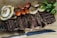 A beautifully arranged plate of sliced grilled steak on a wooden board accompanied by grilled onions, sautéed mushrooms, roasted cherry tomatoes, and fresh parsley. A serrated knife is placed nearby, adding to the rustic presentation.
