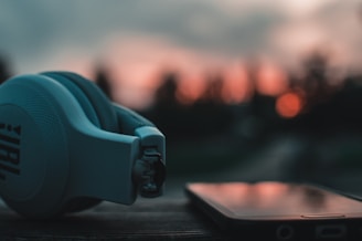 A pair of blue headphones is placed on a wooden surface beside a smartphone. The background is blurred, showing a warm, orange sunset and a soft-focus landscape.