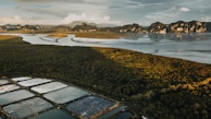 An expansive aerial view captures an intricate landscape featuring a dense, verdant forest bordered by winding waterways. In the foreground, several rectangular fish ponds or salt pans are visible, adding a geometric contrast to the natural surroundings. Rugged mountains rise in the background, under a sky partially filled with clouds.