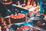 Various types of cakes are displayed on metal trays in a bakery or dessert shop. The cakes are decorated with chocolate or icing, and some have slices cut out, revealing rich, dense textures. The warm, ambient lighting enhances the appeal of the desserts.