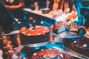 Various types of cakes are displayed on metal trays in a bakery or dessert shop. The cakes are decorated with chocolate or icing, and some have slices cut out, revealing rich, dense textures. The warm, ambient lighting enhances the appeal of the desserts.