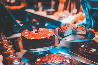 Various types of cakes are displayed on metal trays in a bakery or dessert shop. The cakes are decorated with chocolate or icing, and some have slices cut out, revealing rich, dense textures. The warm, ambient lighting enhances the appeal of the desserts.