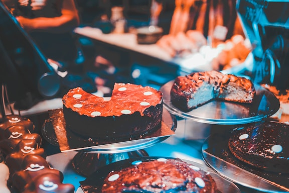 Various types of cakes are displayed on metal trays in a bakery or dessert shop. The cakes are decorated with chocolate or icing, and some have slices cut out, revealing rich, dense textures. The warm, ambient lighting enhances the appeal of the desserts.
