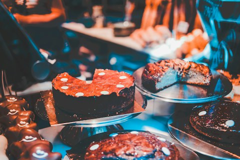 Various types of cakes are displayed on metal trays in a bakery or dessert shop. The cakes are decorated with chocolate or icing, and some have slices cut out, revealing rich, dense textures. The warm, ambient lighting enhances the appeal of the desserts.