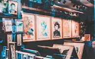 An art gallery display with a variety of abstract and colorful paintings in wooden frames. The walls are covered with artwork, and there are shelves with books and art supplies. Reflections on the glass give a layered, artistic effect.