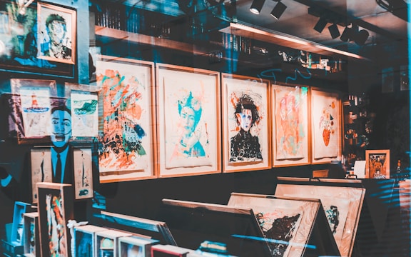 An art gallery display with a variety of abstract and colorful paintings in wooden frames. The walls are covered with artwork, and there are shelves with books and art supplies. Reflections on the glass give a layered, artistic effect.