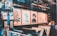 An art gallery display with a variety of abstract and colorful paintings in wooden frames. The walls are covered with artwork, and there are shelves with books and art supplies. Reflections on the glass give a layered, artistic effect.