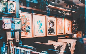An art gallery display with a variety of abstract and colorful paintings in wooden frames. The walls are covered with artwork, and there are shelves with books and art supplies. Reflections on the glass give a layered, artistic effect.