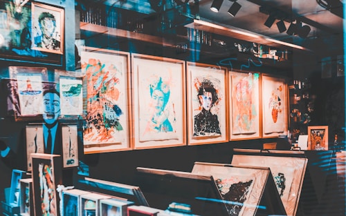 An art gallery display with a variety of abstract and colorful paintings in wooden frames. The walls are covered with artwork, and there are shelves with books and art supplies. Reflections on the glass give a layered, artistic effect.