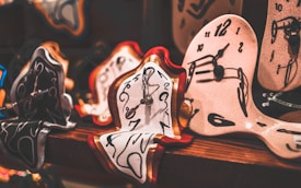 Several stylized clocks with melting designs, reminiscent of surrealist art, are displayed on a wooden surface. The clocks have distorted, fluid shapes and feature prominent black numbers and hands.