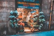 A quaint fruit and vegetable shop with a variety of produce displayed in wooden crates. Numerous bags of fresh greens hang at the entrance, surrounded by stone walls. The shop is bustling with a colorful assortment of fruits and vegetables, showcasing an abundance of natural produce.