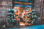A quaint fruit and vegetable shop with a variety of produce displayed in wooden crates. Numerous bags of fresh greens hang at the entrance, surrounded by stone walls. The shop is bustling with a colorful assortment of fruits and vegetables, showcasing an abundance of natural produce.