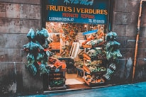 A quaint fruit and vegetable shop with a variety of produce displayed in wooden crates. Numerous bags of fresh greens hang at the entrance, surrounded by stone walls. The shop is bustling with a colorful assortment of fruits and vegetables, showcasing an abundance of natural produce.