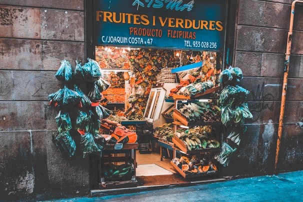 A quaint fruit and vegetable shop with a variety of produce displayed in wooden crates. Numerous bags of fresh greens hang at the entrance, surrounded by stone walls. The shop is bustling with a colorful assortment of fruits and vegetables, showcasing an abundance of natural produce.