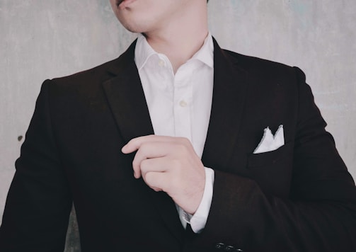 A person is adjusting their black suit jacket, which is worn over a white dress shirt. The hand is lightly touching the jacket, and a white pocket square is neatly tucked into the chest pocket. The background is a neutral, textured wall.