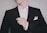 A person is adjusting their black suit jacket, which is worn over a white dress shirt. The hand is lightly touching the jacket, and a white pocket square is neatly tucked into the chest pocket. The background is a neutral, textured wall.