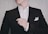 A person is adjusting their black suit jacket, which is worn over a white dress shirt. The hand is lightly touching the jacket, and a white pocket square is neatly tucked into the chest pocket. The background is a neutral, textured wall.