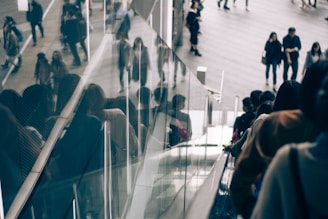 A group of people is using an escalator, moving downwards. The glass panels alongside reflect the images of the individuals, creating a sense of depth and crowd. In the background, more people are walking on a spacious area, giving a busy urban atmosphere.