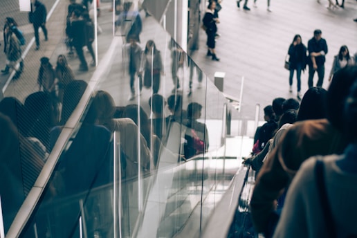 A group of people is using an escalator, moving downwards. The glass panels alongside reflect the images of the individuals, creating a sense of depth and crowd. In the background, more people are walking on a spacious area, giving a busy urban atmosphere.