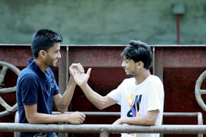 Two young men are engaged in a friendly handshake, smiling at each other. They are standing outdoors near a metal structure.