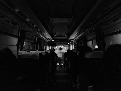 A black and white interior shot of a bus during a nighttime journey. Passengers are seated with their heads silhouetted against the dimly lit ceiling and windows on each side. The aisle leads towards the front where the driver is faintly visible.