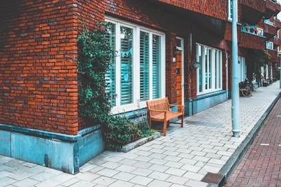 A street view featuring a brick residential building with white-framed windows and a wooden bench on a tiled sidewalk. There is greenery climbing up the side of the building, and a bicycle is partially visible near a lamppost alongside the building.