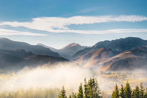 A breathtaking landscape featuring a misty valley surrounded by majestic mountains under a clear, blue sky. The foreground consists of lush green pine trees leading into the fog-covered village below. The mountains display varying shades of grey, brown, and green, illuminated by the soft morning or evening light.