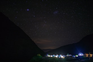 A nighttime scene with a clear starry sky over a campsite. Tents and small buildings are illuminated at the bottom, nestled between dark silhouettes of mountains.