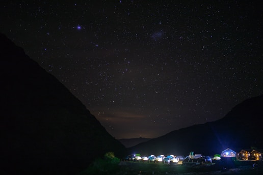 A nighttime scene with a clear starry sky over a campsite. Tents and small buildings are illuminated at the bottom, nestled between dark silhouettes of mountains.