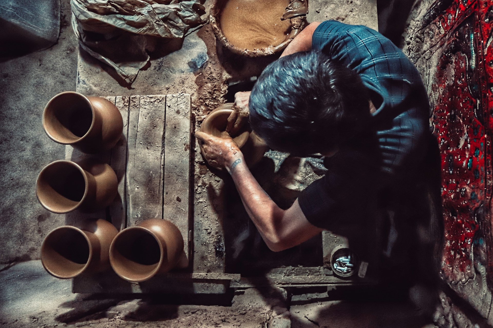 A person is shaping clay on a pottery wheel, surrounded by several finished clay pots. The setting appears rustic with earth tones and a rough-textured environment. The individual is focused on molding the clay, creating a sense of craftsmanship and manual labor.