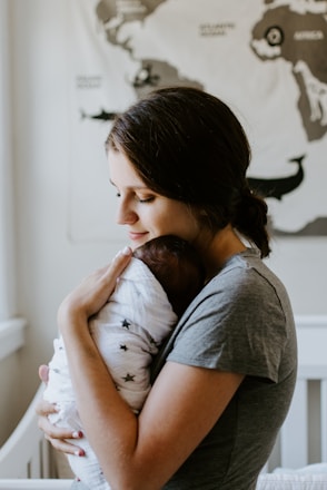 A woman holds a newborn baby who is wrapped in a white blanket with star patterns. Her expression is gentle and tender as she cradles the infant close to her chest. The background features a map on the wall, contributing to a serene setting.