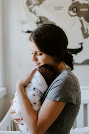 A woman holds a newborn baby who is wrapped in a white blanket with star patterns. Her expression is gentle and tender as she cradles the infant close to her chest. The background features a map on the wall, contributing to a serene setting.