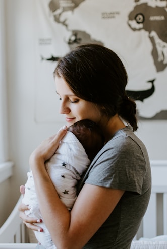 A woman holds a newborn baby who is wrapped in a white blanket with star patterns. Her expression is gentle and tender as she cradles the infant close to her chest. The background features a map on the wall, contributing to a serene setting.