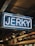 A neon sign displaying the word 'Jerky' in bright white letters against a darker indoor background. Below the sign, there is signage indicating various meat options including beef, pork, and lamb.
