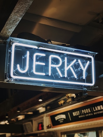 A neon sign displaying the word 'Jerky' in bright white letters against a darker indoor background. Below the sign, there is signage indicating various meat options including beef, pork, and lamb.
