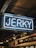 A neon sign displaying the word 'Jerky' in bright white letters against a darker indoor background. Below the sign, there is signage indicating various meat options including beef, pork, and lamb.