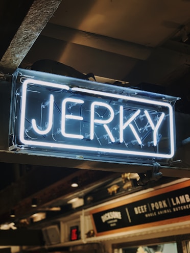 A neon sign displaying the word 'Jerky' in bright white letters against a darker indoor background. Below the sign, there is signage indicating various meat options including beef, pork, and lamb.