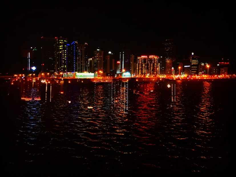 A vibrant city skyline at night with numerous skyscrapers illuminated in various colors. The reflection of these lights shimmers on the water in the foreground, creating a colorful and dynamic scene.