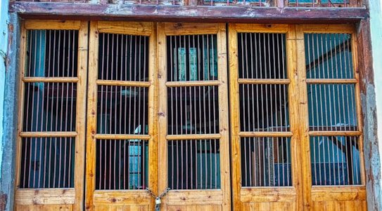 A rustic wooden door featuring vertically aligned metal bars, divided into sections by horizontal wooden frames. The door appears weathered, with visible signs of aging. Some parts of the surrounding wall show chipped paint, adding to the rustic appearance.