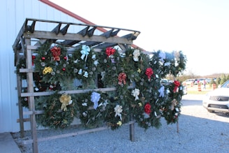 A wooden stand displaying multiple decorative Christmas wreaths adorned with various ornaments such as ribbons, pine cones, and holiday messages. The wreaths are crafted from green foliage and are embellished with bows of different colors including red, gold, and silver. The stand is placed outdoors on a gravel surface next to a white building with a red roof trim. Some parked cars and orange traffic cones are visible in the background.