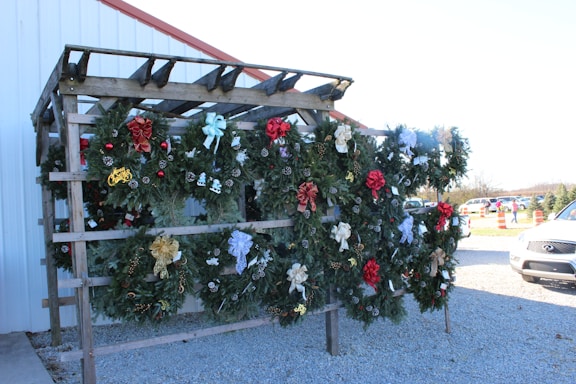 A wooden stand displaying multiple decorative Christmas wreaths adorned with various ornaments such as ribbons, pine cones, and holiday messages. The wreaths are crafted from green foliage and are embellished with bows of different colors including red, gold, and silver. The stand is placed outdoors on a gravel surface next to a white building with a red roof trim. Some parked cars and orange traffic cones are visible in the background.