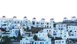 A hillside village composed of whitewashed buildings with blue accents and red-roofed windmills sparsely distributed among the houses. The architecture is traditional and simple, featuring flat roofs and minimalistic design. The overall atmosphere is Mediterranean, likely depicting a scenic island setting.